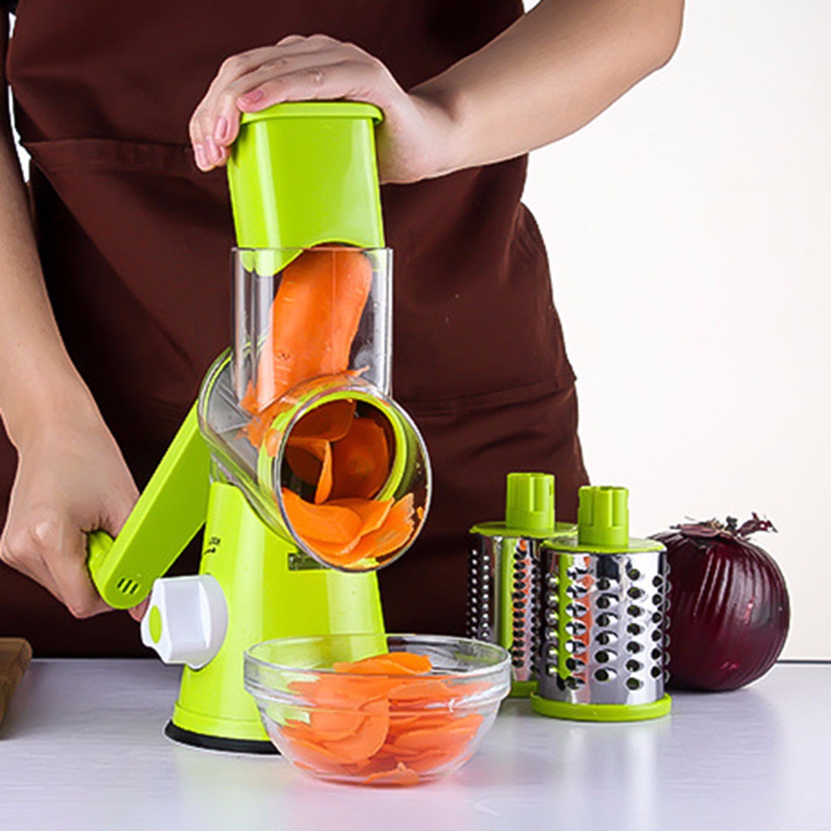 Person using a green manual vegetable slicer to slice carrots, with a bowl of sliced carrots and additional grating attachments on a white surface.