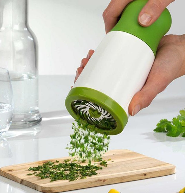Hand using a green and white herb chopper on a wooden cutting board with chopped herbs.