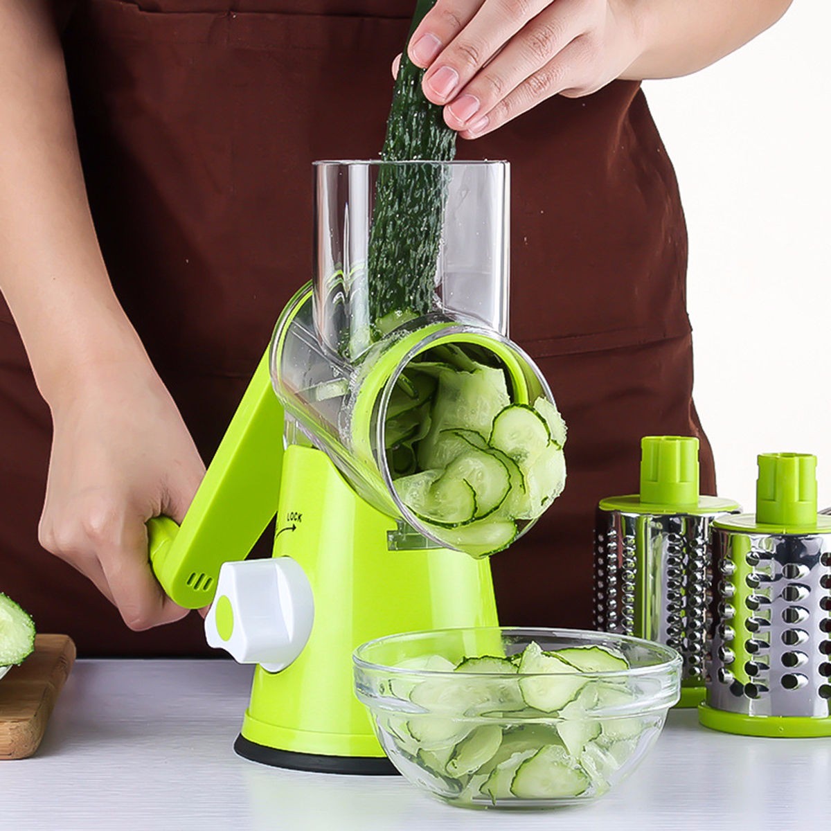 Person using a green manual vegetable slicer to slice cucumbers, with sliced cucumbers in a bowl and additional slicer parts in the background.